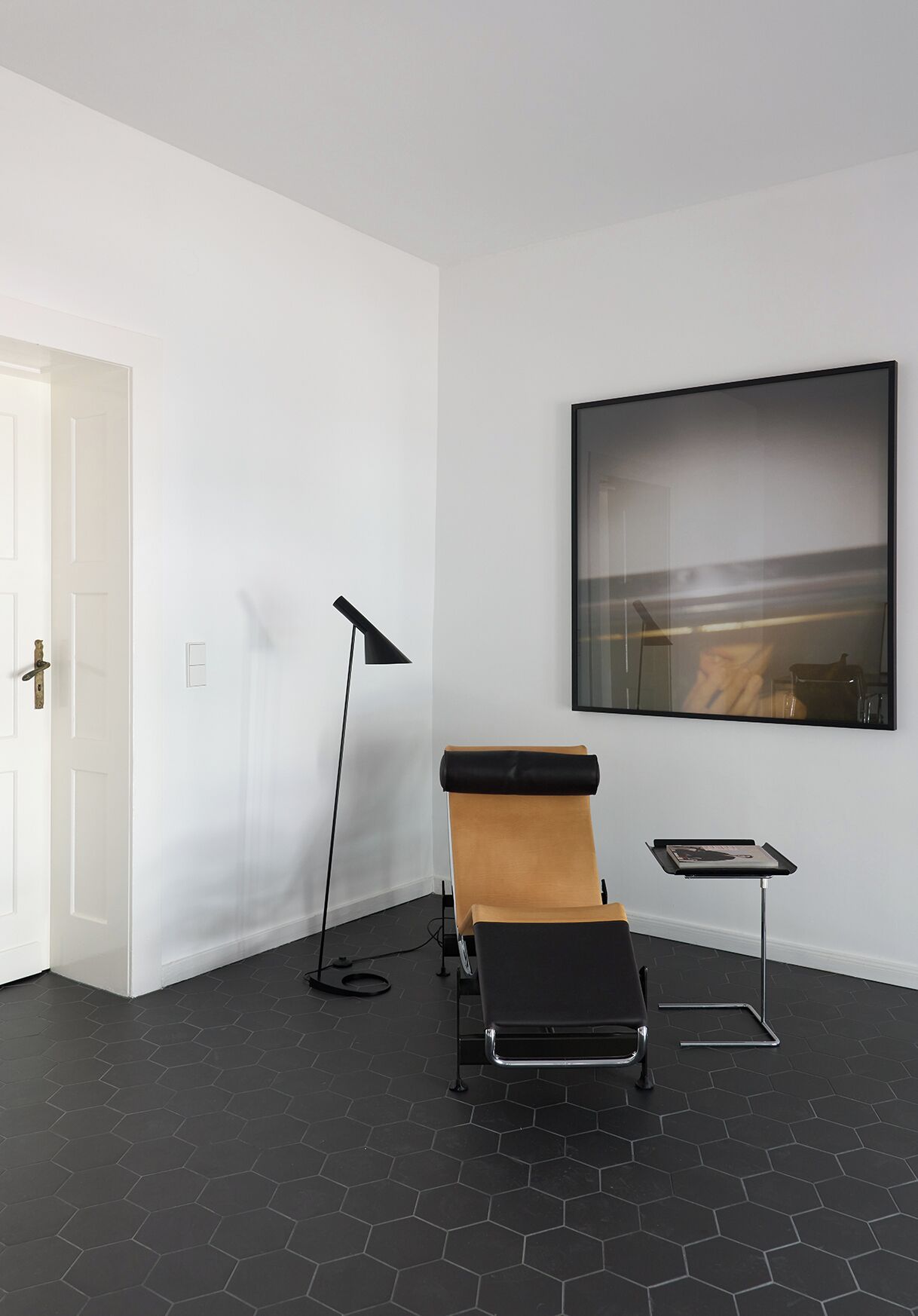 A room with a black hexagonal floor, a white door, a black floor lamp, a massage table, and a framed picture on the wall.