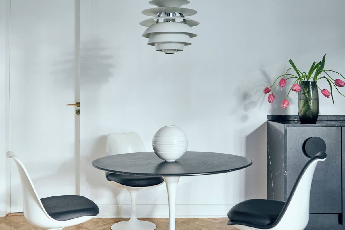 A dining area with a round table, two white chairs with black cushions, a modern pendant light, a vase with pink flowers, and a cabinet with a round mirror.