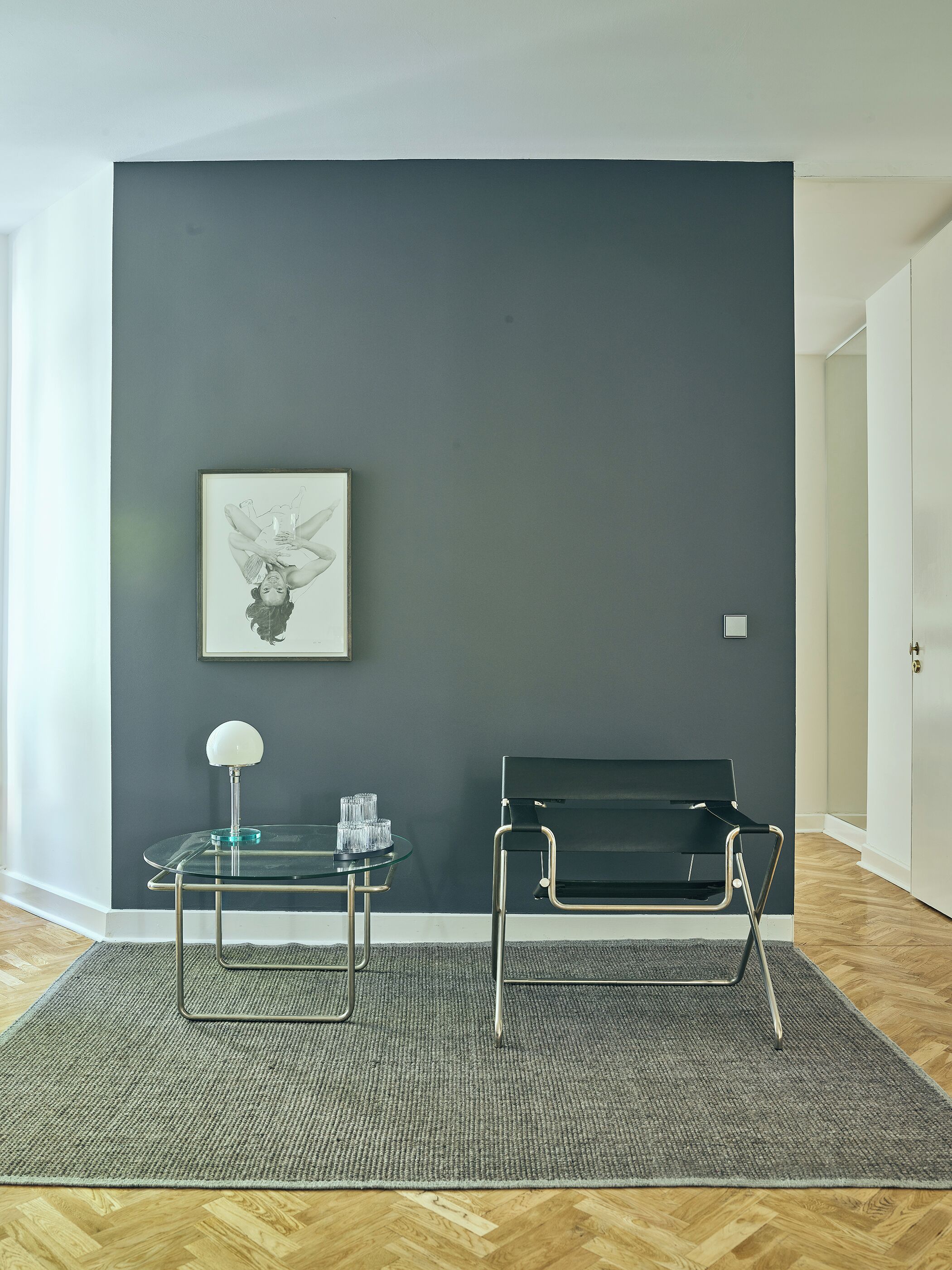 Een moderne kamer met een donkergrijze wand, een glazen tafel met een lamp, een zwarte bank en een ingelijst schilderij.