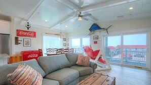 Ein Wohnzimmer mit grauem Ecksofa, einem Holztisch und einem roten Coca-Cola Kühlschrank. Ein großes Fenster bietet Blick auf ein Gebäude und einen Balkon.
