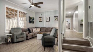 A living room with a grey sofa set, a grey armchair, a coffee table, and a ceiling fan. There are framed pictures on the wall and a window with blinds.