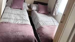 Ein gemütliches Schlafzimmer mit zwei Betten, jeweils mit Kopfstütze und passender Kopfkissen. Darüber befindet sich ein Holzregal, und ein Fenster mit Vorhang.