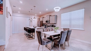 A modern kitchen with a central island, bar stools, and a dining table with chairs. - Game Room & Private Pool! Windsor Cay 5BR Home (Clermont)