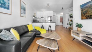 Un salon moderne avec un canapé en cuir noir, une table basse en bois et une cuisine en arrière-plan.