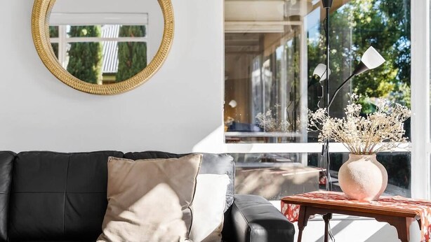 Un salon moderne avec un canapé en cuir noir, un miroir rond et un vase de fleurs posé sur une petite table.