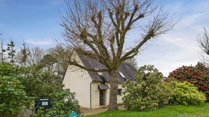 Een huis met een grote boom ervoor, omringd door groen en een heldere lucht.