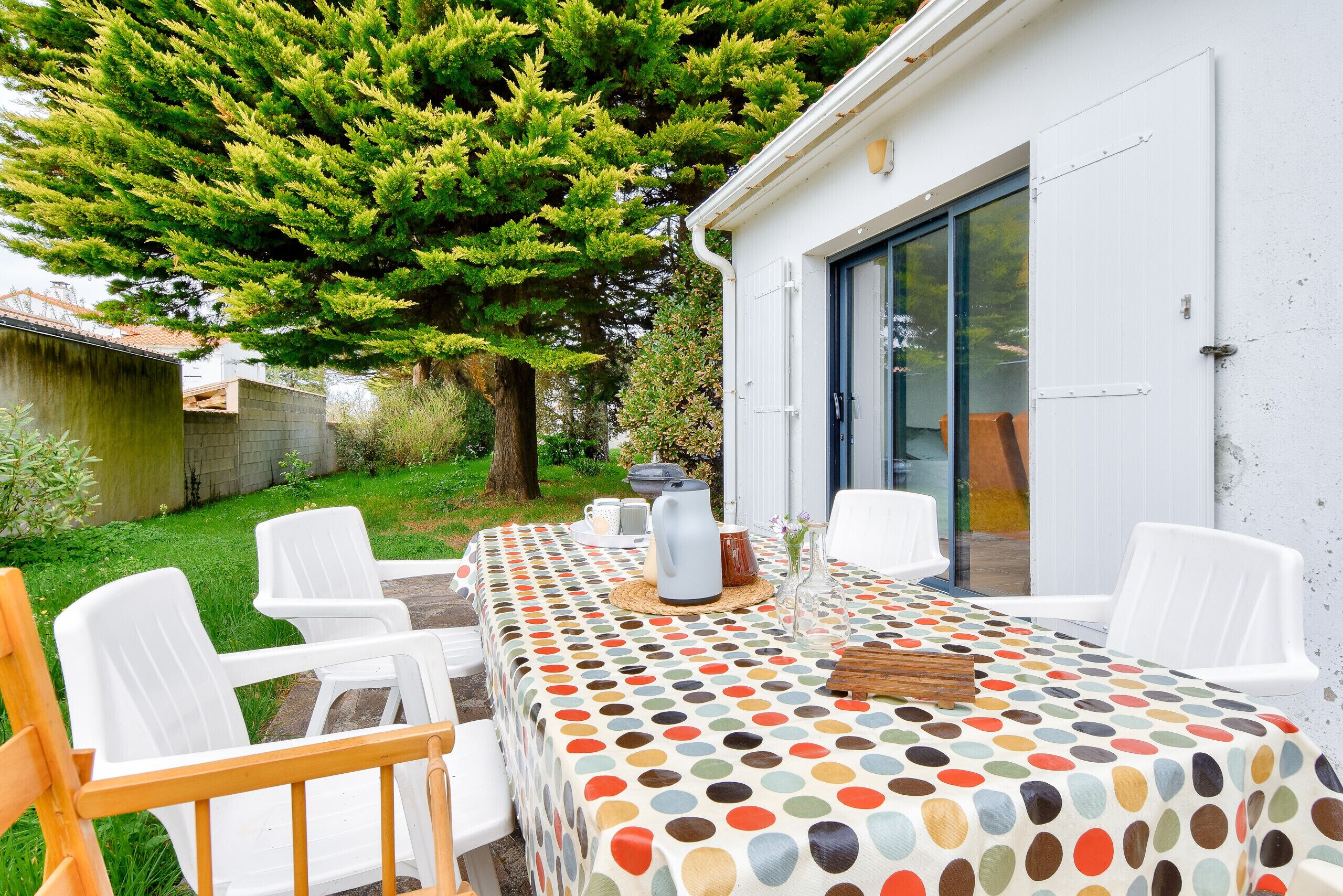 Un espace extérieur aménagé pour les repas, avec une table recouverte d’une nappe à pois, entourée de chaises en plastique blanches, le tout se fondant dans un décor composé d’un arbre verdoyant et d’un bâtiment blanc doté d’une porte-fenêtre coulissante.