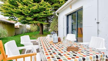 An outdoor dining area with a table covered in a polka dot tablecloth, surrounded by white plastic chairs, set against a backdrop of a lush green tree and a white building with a sliding glass door.