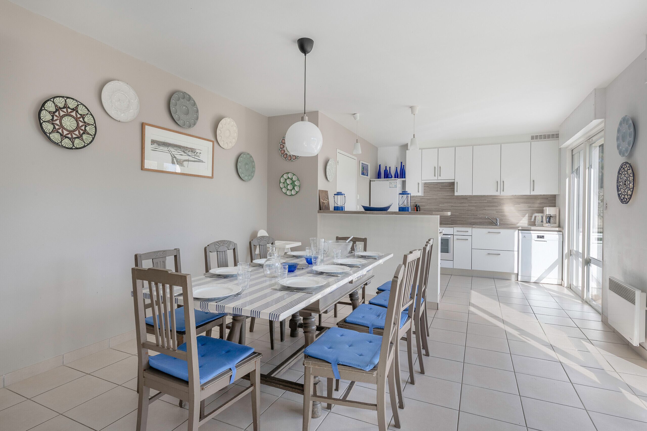 Een eethoek met een lange tafel en stoelen, een keuken op de achtergrond, en decoratieve borden aan de wanden.