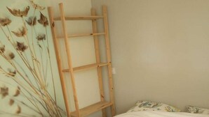 Un lit bien fait, avec une serviette verte et deux tapis de bain jaunes, une échelle en bois appuyée contre le mur et des fleurs séchées accrochées au mur.