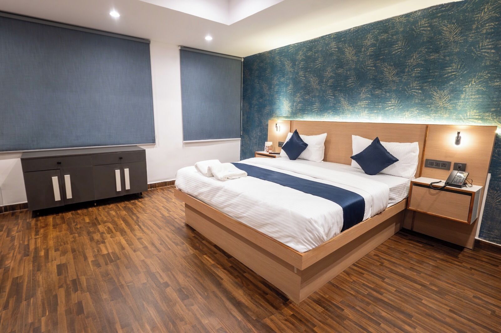 Een moderne hotelkamer met een groot bed, een houten hoofdbord, nachtkastjes met lampen, een kledingkast en een groot raam met jaloezieën.