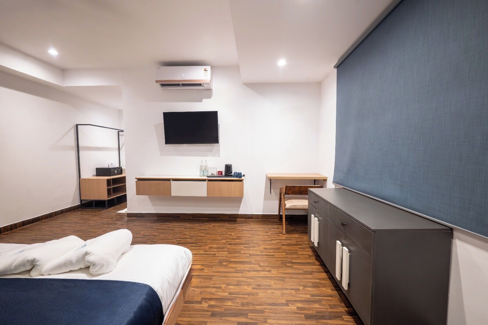 A modern hotel room with a bed, a desk, a chair, a flat-screen TV, a wardrobe, and an air conditioning unit.