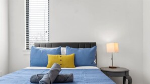 A neatly made bed with blue bedding and a yellow pillow, a bedside lamp, and a window with blinds.
