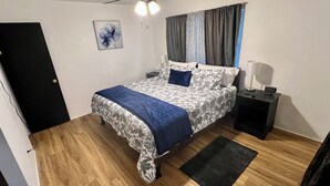 Une chambre à coucher avec un lit, une table de chevet, une lampe, une porte et une fenêtre avec des rideaux.