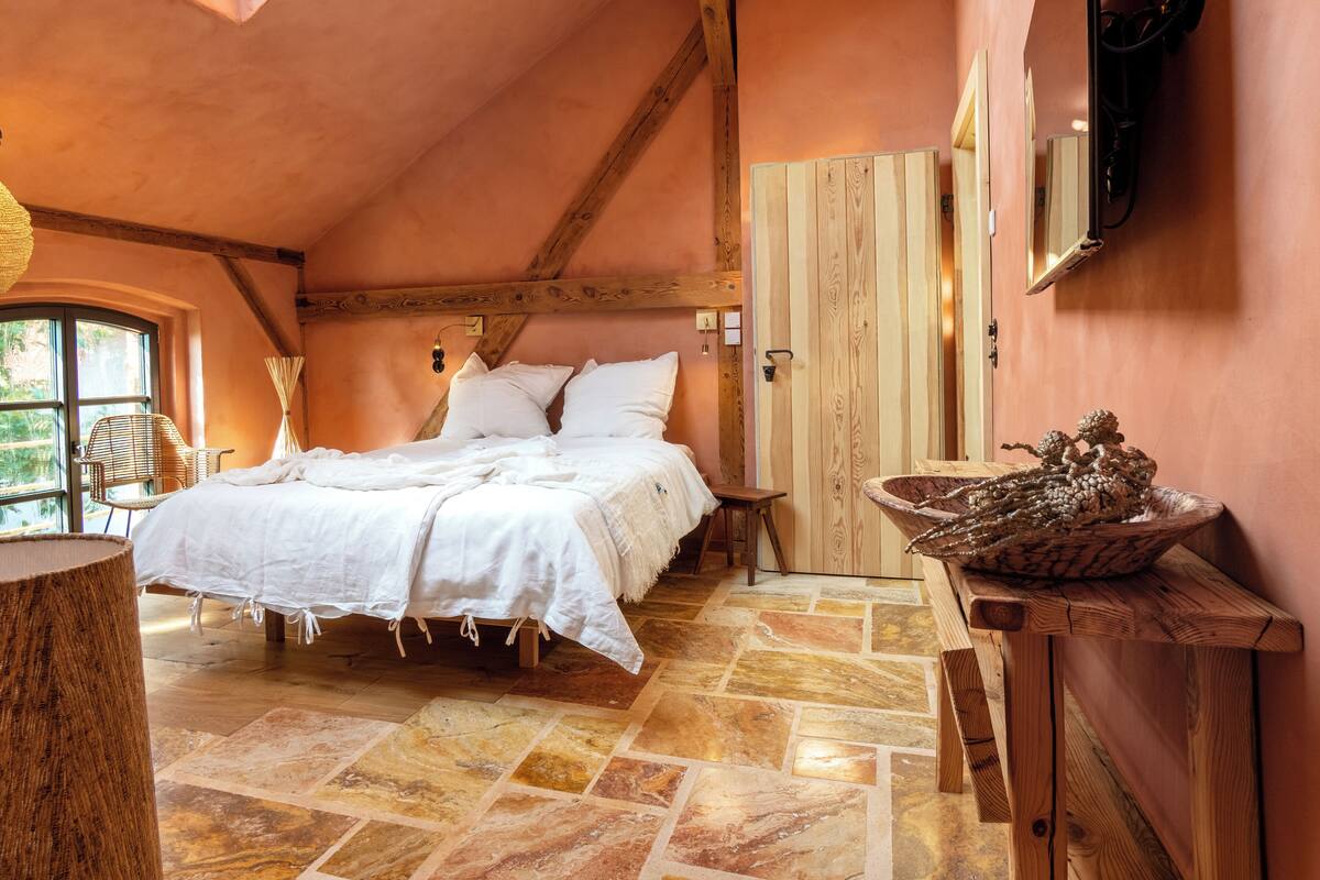 Un dormitorio con cama, vigas de madera, suelo de piedra, puerta de madera y televisor montado en la pared.