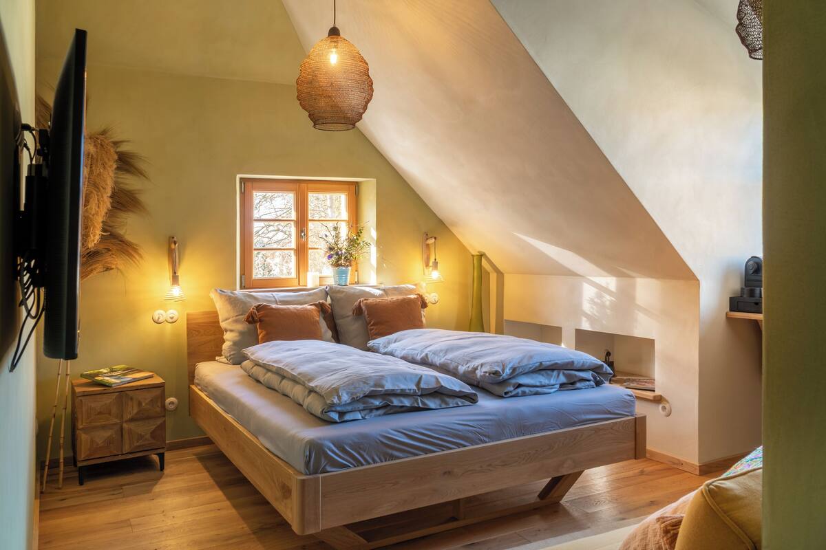 Un dormitorio con techo inclinado, una cama con ropa de cama azul, una mesita de noche, una lámpara y una ventana con cortinas.