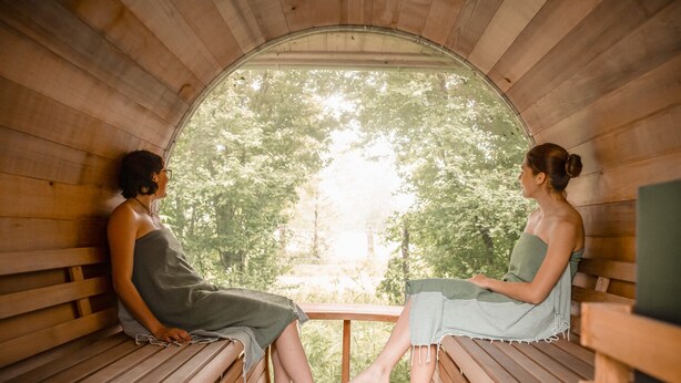 Twee personen zitten op houten banken in een sauna, met uitzicht op bomen door een halfrond raam.
