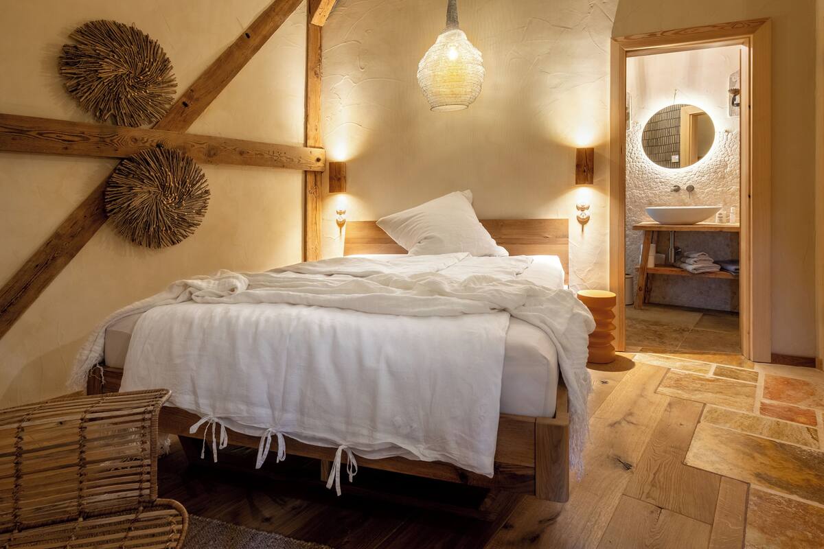 Un dormitorio con cama de madera, ropa de cama blanca, un adorno de pared tejido, una lámpara colgante y vista al baño.