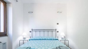 Una cama individual con estructura metálica, dos mesitas de noche con lámparas y una ventana a la izquierda.