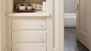 Un pasillo con una puerta color crema, un estante con plantas y un vistazo a un baño con bañera.