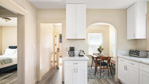 Cocina compacta con armarios blancos, comedor con mesa y sillas, y un vano visible que da a otra habitación.