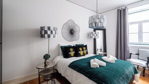 A modern hotel room with a neatly made bed, a small side table with a plant, a chair, and a large window with curtains. - Rosemary Apartment, Campo de Ourique, Lisbon (Lisboa)