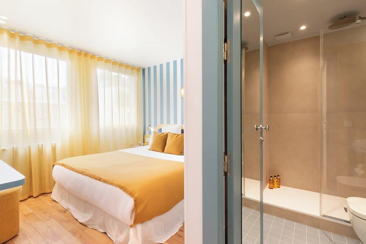 A bedroom with a bed, a bedside table, and a window with curtains. The room has a light-colored floor and walls.