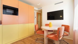 A modern kitchen with orange and yellow cabinets, a white dining table with four orange chairs, and a flat-screen TV mounted on the wall.