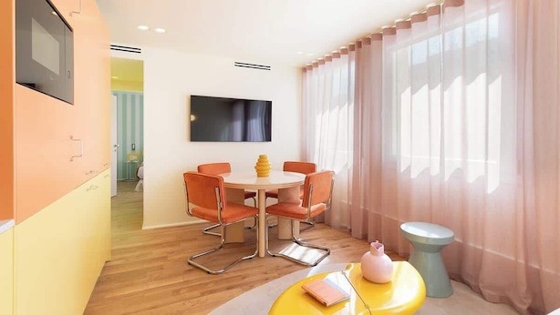 A modern hotel room with a round dining table, orange chairs, a yellow ottoman, and a TV mounted on the wall.