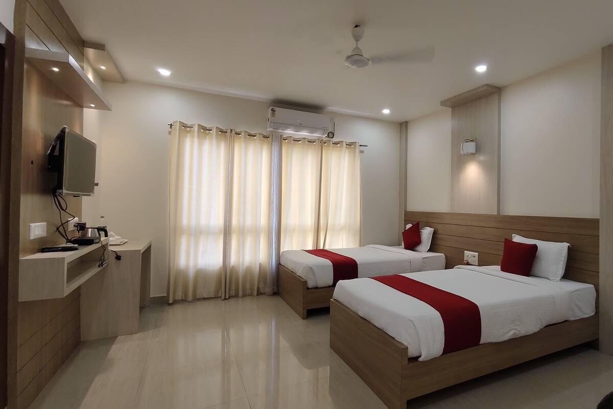 A hotel room with two beds, a TV mounted on the wall, a ceiling fan, and a window with curtains.