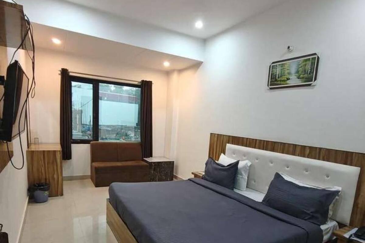 A modern bedroom with a large bed, a wooden headboard, a nightstand, a TV mounted on the wall, and a window with curtains.