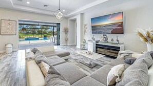 A spacious living room with a large TV, comfortable seating, and a view of a pool and outdoor area. - Esplanade by the Islands (Naples)