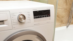 Lavadora de carga frontal con panel de control y puerta de cristal, ubicada en un baño con piso de baldosas.