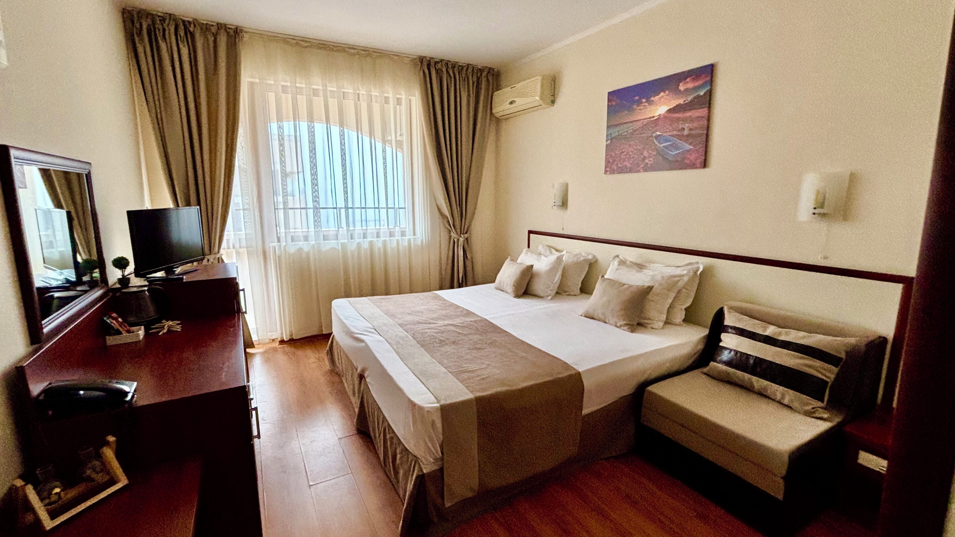 A hotel room with a bed, a desk, a chair, a television, and a painting on the wall.