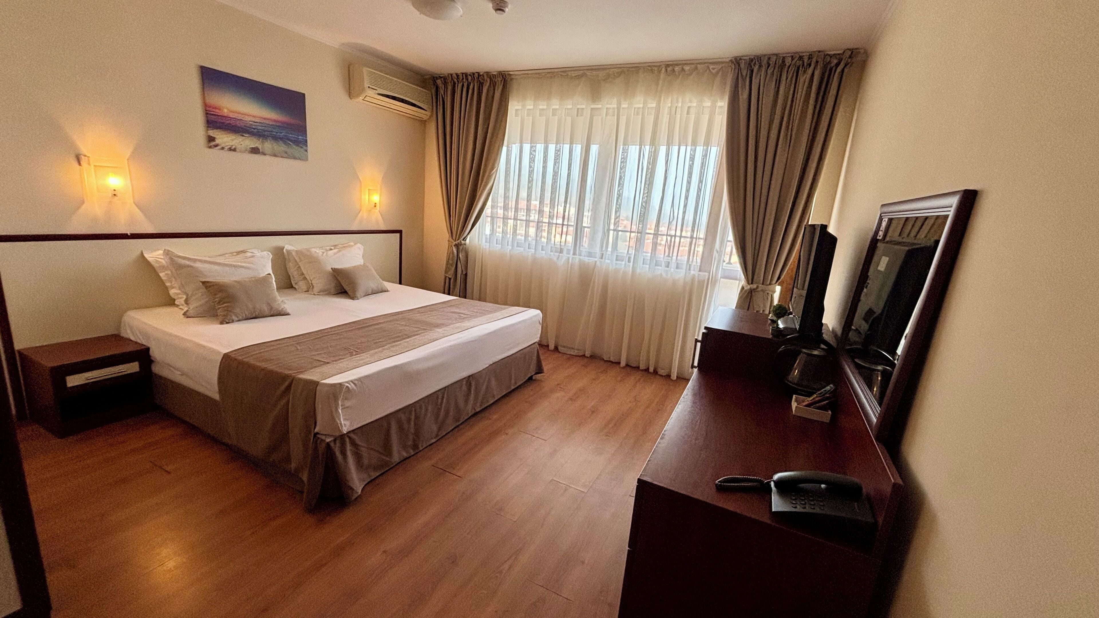 Ein Hotelzimmer mit Bett, Nachttischen, einem Schreibtisch mit Telefon, einem Spiegel und einem fenster mit Vorhängen.
