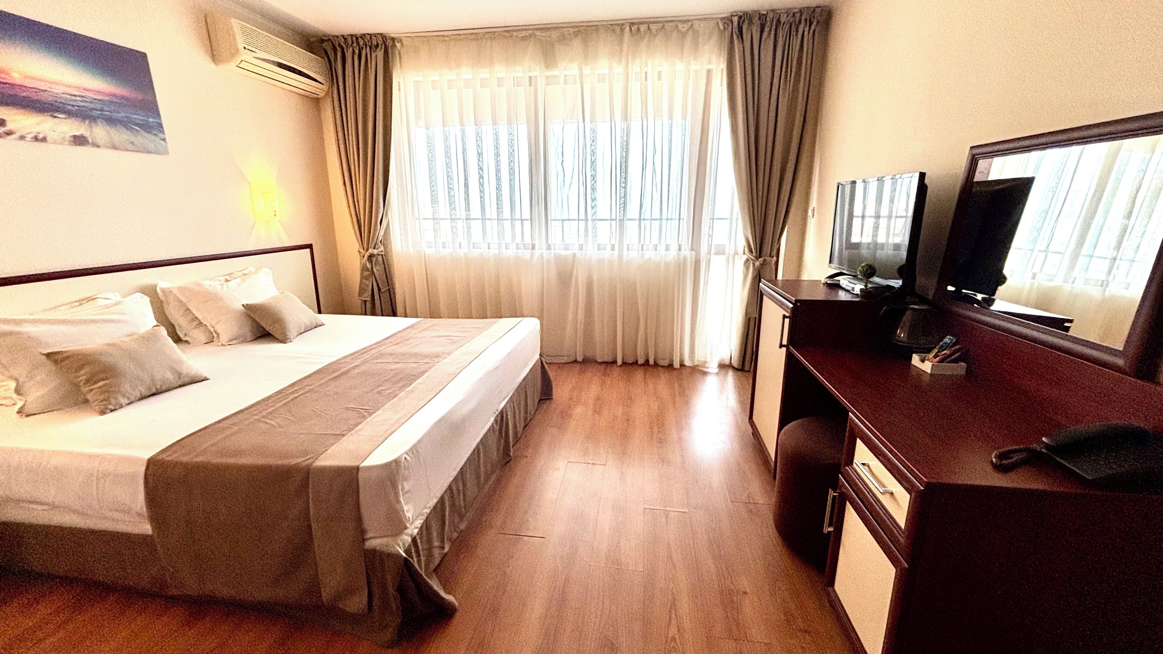 Quarto de hotel com cama, escrivaninha, computador, televisão e janela com cortinas.