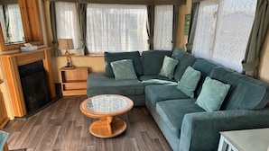 A cozy living area with a sectional sofa, a round wooden coffee table, and a fireplace. - Family Caravan - Sleeps 6 - Pets Allowed - Pool (King's Lynn)