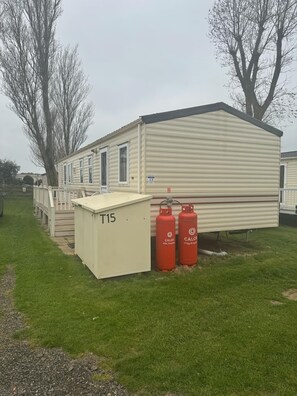 Una casa móvil con techo plano, un porche cubierto y dos tanques de propano etiquetados como 'CALOR'.