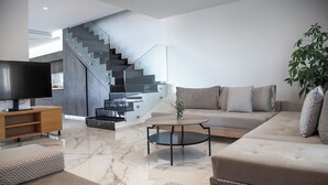 Un salón moderno con sofá, mesa de centro y televisor sobre un mueble de madera. Se ve una escalera con barandilla de vidrio al fondo.