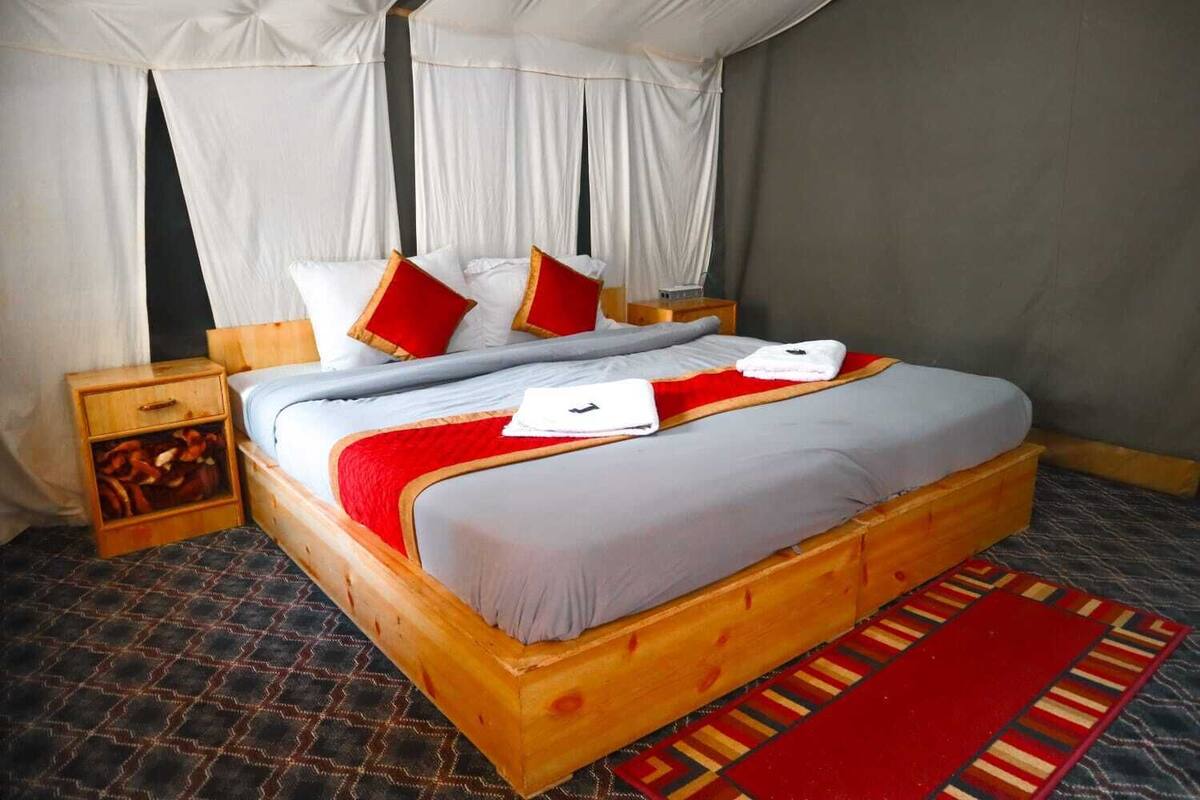 Ein Bett mit Holzrahmen, weißer Bettwäsche, roten Kissen und einem Teppich mit roten und goldenen Streifen.