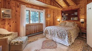 Un dormitorio de cabaña de madera acogedor con una cama grande, una estantería, una silla de mimbre y una ventana con cortinas.