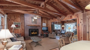 Interior acogedor de una cabaña de madera con chimenea, vigas de madera, televisor y asientos cómodos.