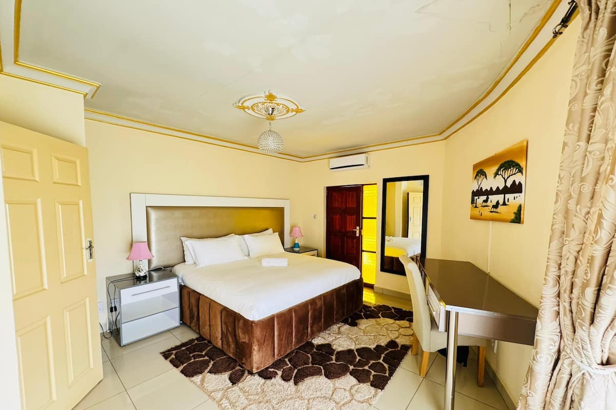 A hotel room with a bed, bedside table, lamp, desk, chair, and a painting on the wall.