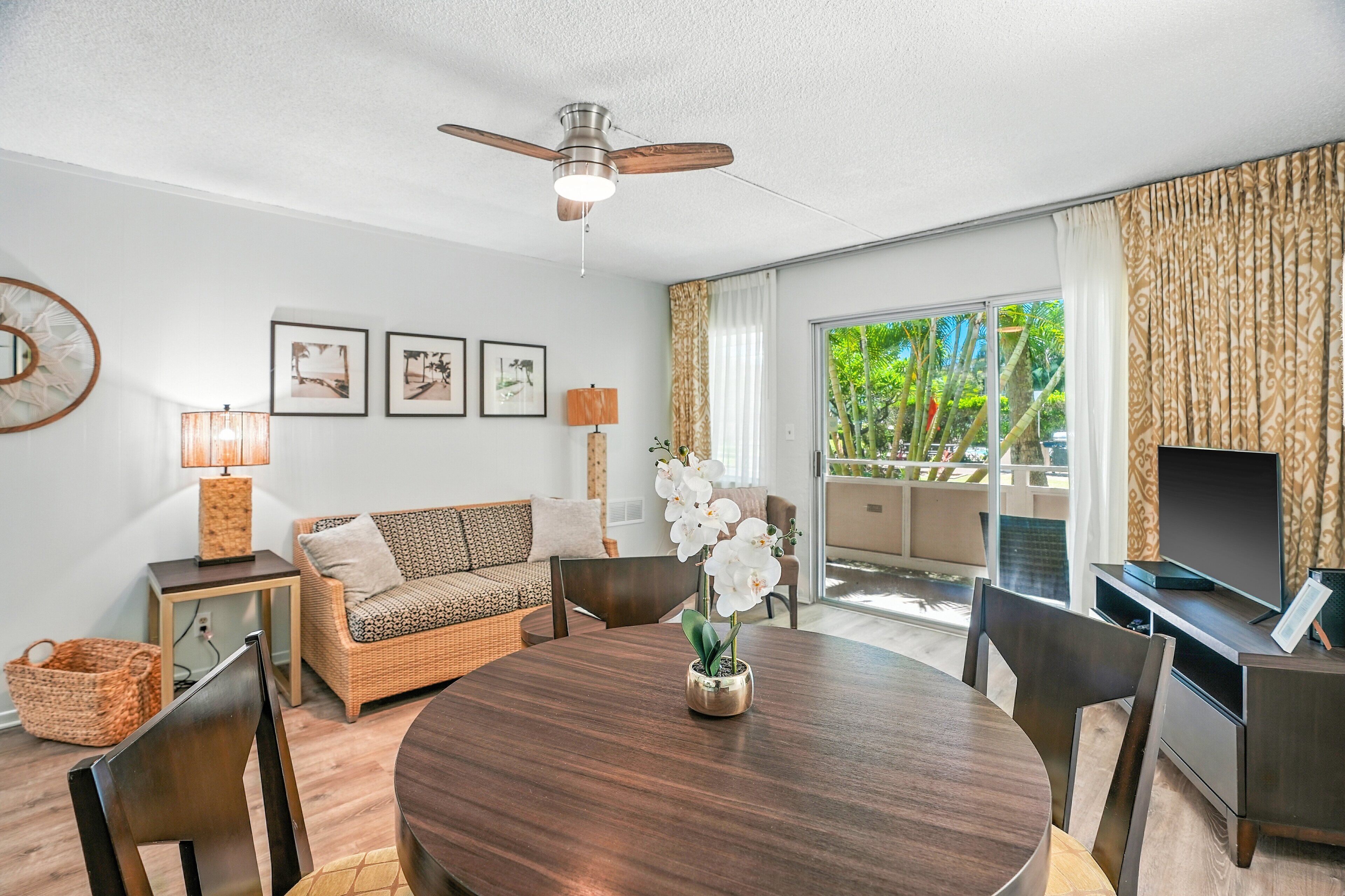 A modern living room with a round wooden dining table, wicker sofa, and a ceiling fan.