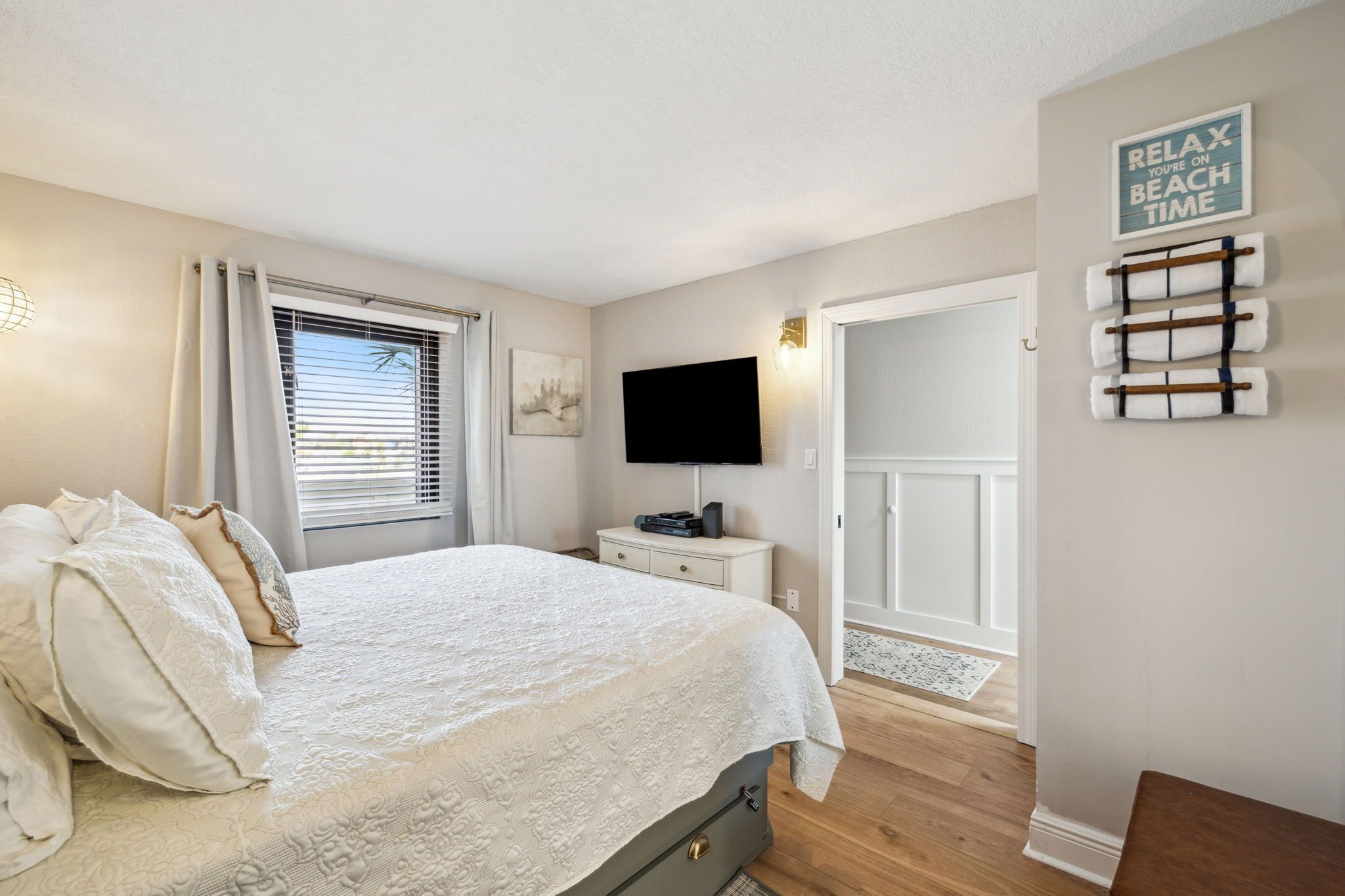 A bedroom with a bed, a TV, a window with curtains, a wall-mounted towel rack, and a door leading to another room.