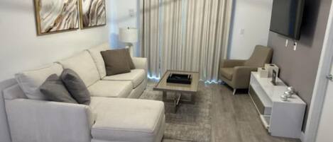 A modern living room with a sofa, armchair, coffee table, and a flat-screen TV mounted on the wall.