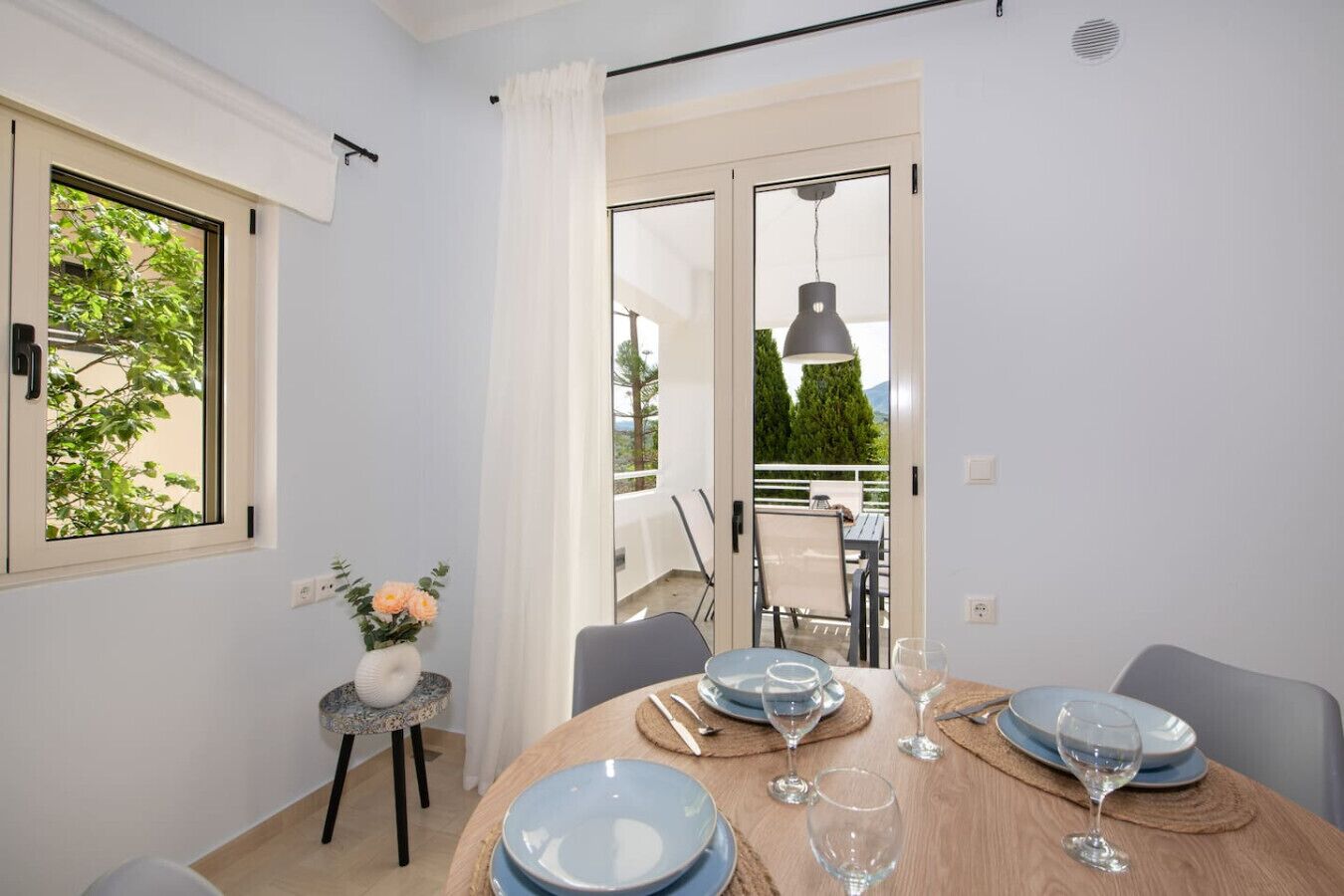 A dining area with a round table set for four, a small side table with a vase of flowers, and a view of the outdoor balcony through the window.