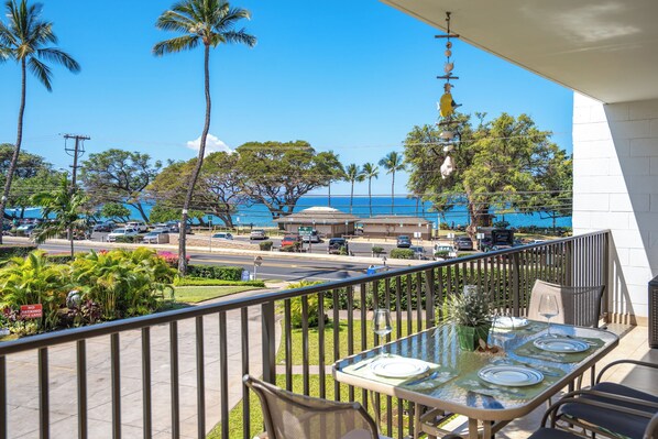 A balcony with a table set for dining, overlooking a street with cars and palm trees, and a view of the ocean.