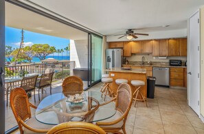 A modern kitchen with wooden cabinets, a refrigerator, and a countertop. There is a dining area with wicker chairs and a glass table. The room has a sliding glass door leading to a balcony with a view of the ocean.