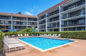 A hotel with a swimming pool surrounded by lounge chairs and umbrellas, multiple balconies, and a clear blue sky.